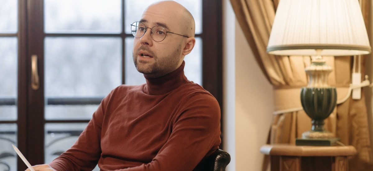 Bald man in glasses speaking while seated indoors, with a warm lamp and curtains in the background.
