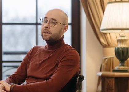 Bald man in glasses speaking while seated indoors, with a warm lamp and curtains in the background.