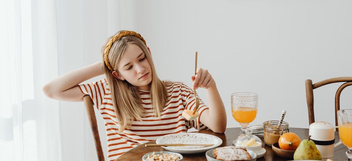 Teen girl in striped shirt at breakfast table with fruits, bread, and juice. Casual morning setting.